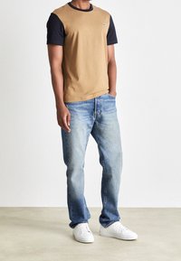 Man står med händerna i fickorna, iklädd en tvåfärgad kortärmad skjorta i tan och svart, ljusblå jeans och vita sneakers på en neutral golv.