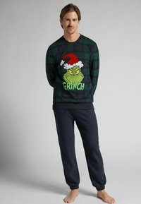 Pánská mikina s kostkovaným vzorem v zelené barvě, která má velký grafický motiv Grinche a text. Vytvořena ve spojení s tmavě námořnickými tepláky, které mají uvolněný střih.