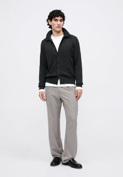 Uomo che indossa un cardigan grigio scuro con bottoni sopra una camicia bianca, pantaloni grigio chiaro e mocassini neri, in piedi su uno sfondo bianco.