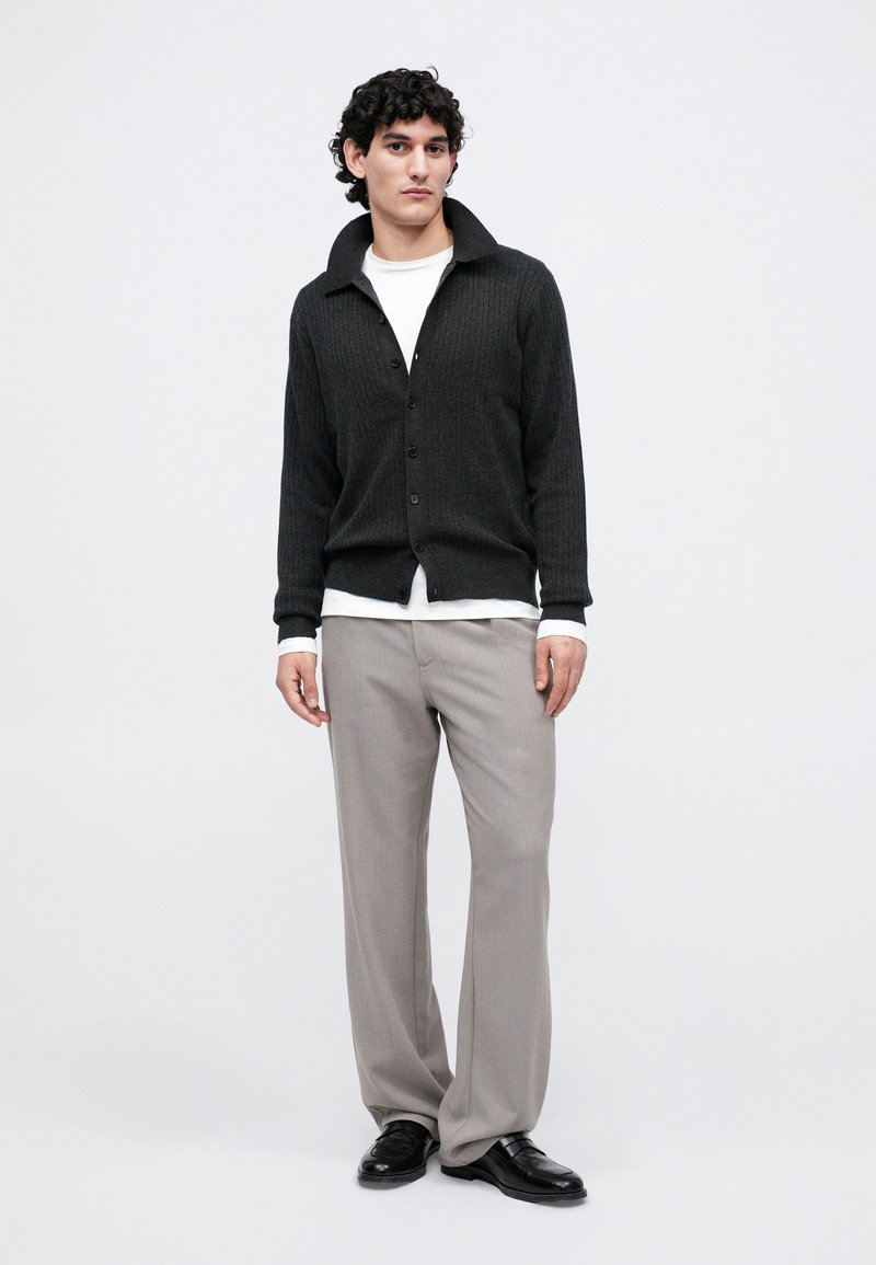 Man wearing a dark gray buttoned cardigan over a white shirt, light gray trousers, and black loafers, standing against a white background.