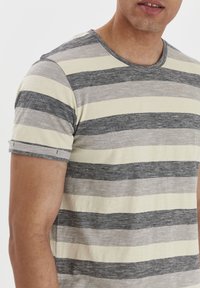 Camiseta de manga corta a rayas que presenta colores gris y crema alternantes, con un cuello redondo y mangas enrolladas. Textura de tela suave.