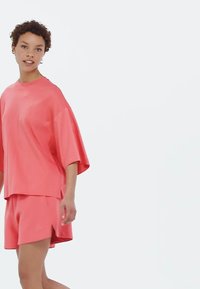 Korálovo ružový športový set pozostávajúci z voľného, krátkeho rukávu a zodpovedajúcich šortiek, vyrobený z hladkej, ľahkej látky.