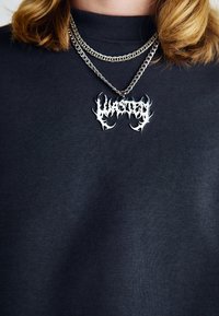 Maglione nero a coste con collo alto e catene d'argento stratificate, caratterizzato da un ciondolo di testo unico e frastagliato che recita "Wasted."