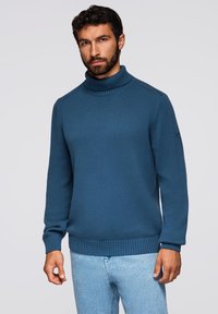 Blauer Strickpullover mit hohem Rollkragen, gerippten Bündchen und Saum. Der Stoff hat ein strukturiertes Muster und ist mit einem Logo am Ärmel versehen.
