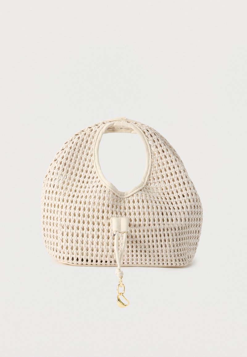 Τσάντα χειρός σε κρεμ απόχρωση με στρογγυλεμένες επάνω λαβές και ένα χρυσό κρεμαστό διακοσμητικό που κρέμεται από το μπροστινό κορδόνι.