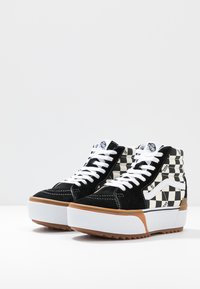 Zapatillas de caña alta en blanco y negro con diseño de tablero de ajedrez, parte superior de ante, cordones blancos y suela de goma con una base marrón texturizada.