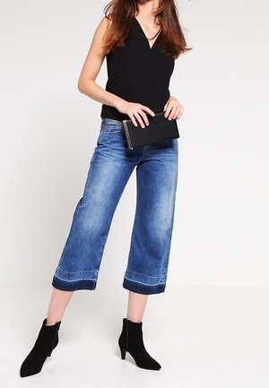 Femme portant un débardeur noir, un jean bleu raccourci, des bottines noires, tenant une pochette noire sur un fond clair uni.