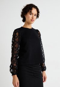 Svart topp med solid kropp og gjennomsiktige, puffede mesh-ermer med blomstermotiv. Teksturert skjørt med rynkedetaljer.