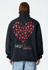 Personne portant une veste polaire noire avec des cœurs rouges brodés formant un grand cœur et le texte « SWEET heart » dans le dos.