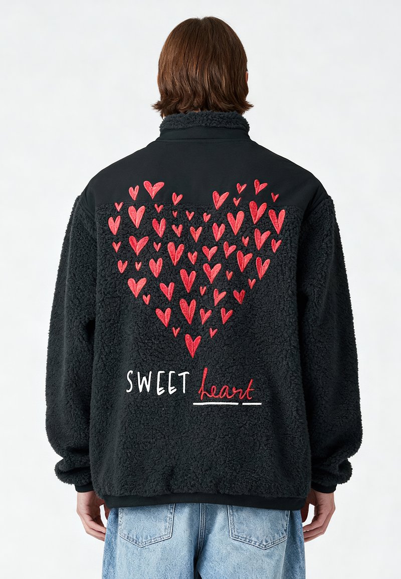 Personne portant une veste polaire noire avec des cœurs rouges brodés formant un grand cœur et le texte « SWEET heart » dans le dos.