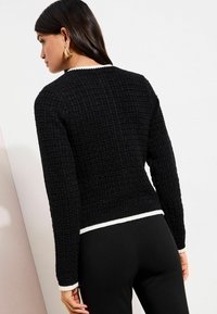 Naine, kes kannab musta tekstuuriga kampsunit valge äärisega krae, varrukaotste ja ääre juures, seljas mustad püksid, seisab ühevärvilise tausta ees.