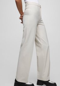 Pantalon beige clair à jambes larges avec surpiqûres contrastantes, taille haute et fermeture éclair, associé à des chaussures à plateforme noires.