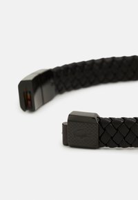 Bracelet en cuir tressé noir avec fermoir métallique carré. Le fermoir présente un motif texturé et un détail de logo gravé. Finition lisse dans l'ensemble.