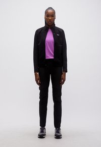 Γυναίκα που στέκεται προς τα εμπρός φορώντας μαύρο σακάκι, μωβ πουκάμισο, μαύρο παντελόνι και παπούτσια πεζοπορίας σε απλό φόντο.