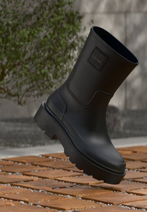 Botte noire Calvin Klein à semelle épaisse flottant au-dessus de lattes en bois et de petits cailloux blancs en plein air près de plantes vertes.