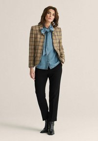 Rutet blazer i brun og beige, parret med en blå denimskjorte og sorte skræddersyede bukser. Sorte ankelstøvler fuldender looket.