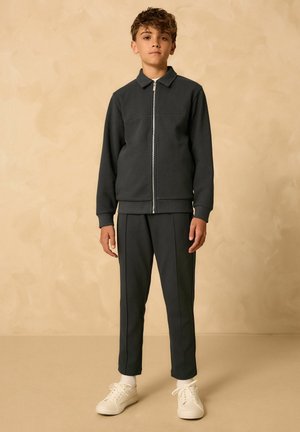 Mørkegrå zip-up jakke med krave og patch-lomme, parret med matchende slim-fit bukser med sidelister. Hvide sneakers fuldender looket.