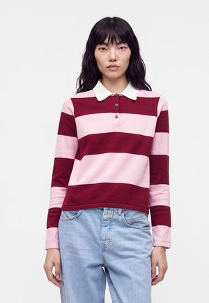 Kvinde iført en langærmet polo med brede horisontale striber i pink og bordeaux samt lyseblå jeans, stående foran en ensfarvet baggrund.