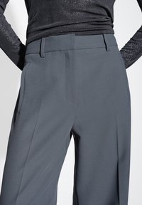 Pantaloni grigi su misura con una texture liscia, dotati di tasche laterali e una vita piatta. Arricchiti da pieghe sottili per un tocco di dettaglio in più.