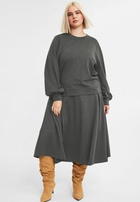 Kobieta z blond bobem, ubrana w ciemnoszary top z długim rękawem i dopasowaną midi spódnicę, w luźnych, beżowych zamszowych botkach.