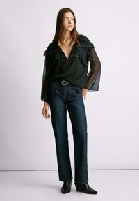 Blusa negra semitransparente con acentos de volantes, combinada con jeans de mezclilla oscuros de cintura alta y botas de tacón negro hasta el tobillo. Detalles de estilo sencillos prominentes.