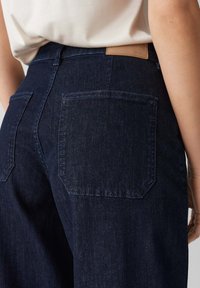 Jeans in denim blu scuro con vita alta, due tasche posteriori e un'etichetta in pelle sul retro. Il tessuto ha una superficie liscia.