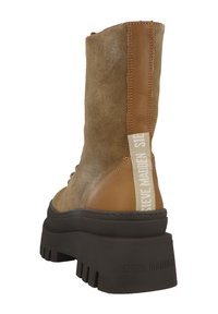 Botín de ante marrón con detalles en cuero marrón claro, suela gruesa de goma negra y una etiqueta de tela con marca en el costado. Diseño robusto.