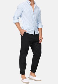 Chemise à boutons à rayures bleu clair et blanc, pantalon noir fuselé et chaussures blanches à enfiler, montrant une coupe décontractée avec les manches retroussées.