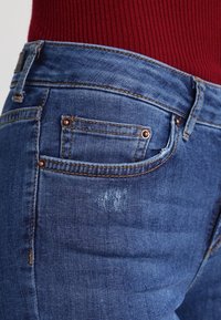 Sininen farkkuhousu klassisella muotoilulla, jossa on takatasku kuparin niiteillä, hienovarainen kuluminen ja teksturoidut vyötärö.