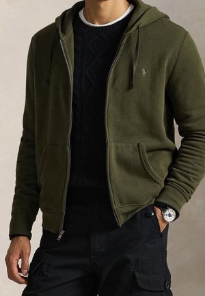 Mann trägt einen olivgrünen Kapuzenpullover mit Reißverschluss über einem schwarzen strukturierten Pullover, schwarze Cargohosen und eine Armbanduhr mit weißem Zifferblatt.