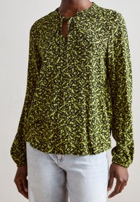 Haut noir avec un motif floral vert, tissu léger, manches longues, col rond avec une ouverture en goutte et une fermeture à lien, coupe décontractée.