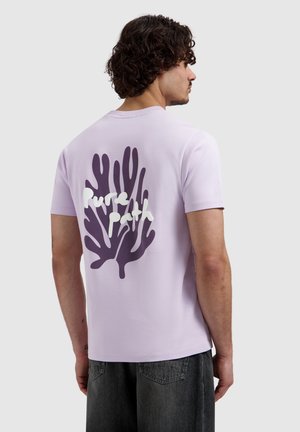 Man met krullend haar draagt een lichtpaarse T-shirt met een donkerpaarse koraalprint en witte tekst "Pure Path" op de rug.