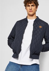 Jachetă bomber matlasată de culoare bleumarin, cu manșete și guler ribbați, având un fermoar frontal și un mic patch cu logo brun pe piept.