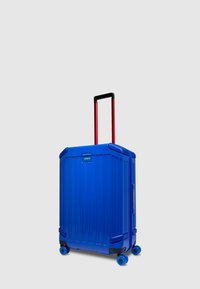 Valigia rigida blu con scanalature verticali, due ruote blu e manico rosso retrattile. Presenta un'etichetta con logo frontale per l'identificazione.