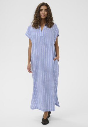 Woman with curly hair wearing a blue and white vertical striped long dress and black woven shoes, standing with one hand in pocket.