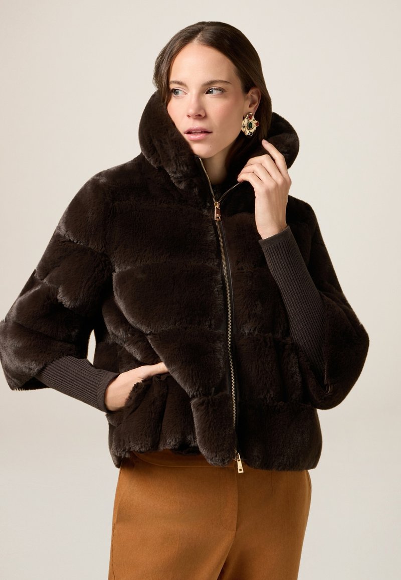 Brązowa kurtka z sztucznego futra z wysokim kołnierzem, suwakiem i szerokimi rękawami. Posiada poziome szwy i miękką, teksturowaną powierzchnię.