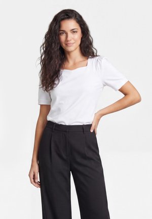 Vrouw met lang, golvend haar draagt een wit top met vierkante hals en korte mouwen en een zwarte broek met hoge taille, staand met één hand op haar heup.