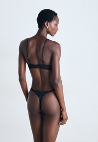 Femme avec les cheveux courts portant un soutien-gorge en dentelle noire et un string, se tenant de profil contre un fond clair et uni.