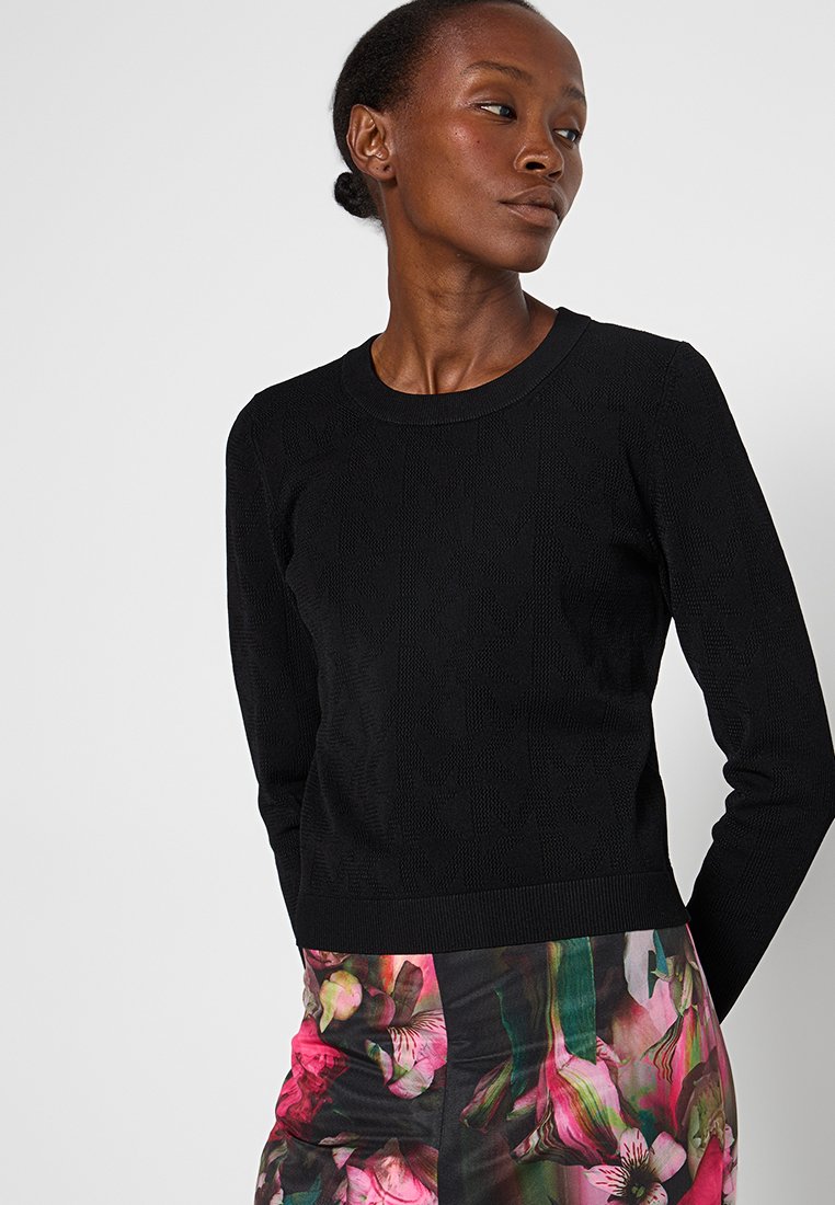 Donna che indossa una maglietta nera a maniche lunghe con texture e una gonna con una vivace stampa floreale rosa e verde, su uno sfondo chiaro uniforme.