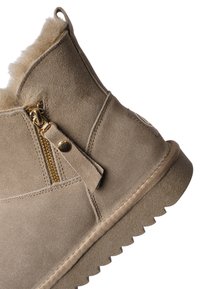 Beiger Wildleder-Winterstiefel mit seitlichem goldfarbenem Reißverschluss, Zuglasche, Fellfutter und strukturiertem Gummisohlenprofil, schräg gestellt, um die Außenseite zu zeigen.