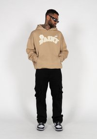 Sweatshirt beige à capuche avec le graphisme blanc "SAINT", associé à un pantalon cargo noir avec plusieurs poches et des baskets noires avec des accents blancs.