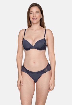 Mujer con el pelo largo y castaño que lleva un conjunto a juego de sujetador y ropa interior de encaje azul oscuro, de pie y sonriendo frente a un fondo liso.
