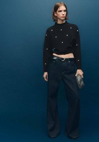 Pullover nero cropped con perline argentate, abbinato a jeans in denim scuri a vita alta. La modella tiene una clutch metallica, in piedi contro uno sfondo blu.