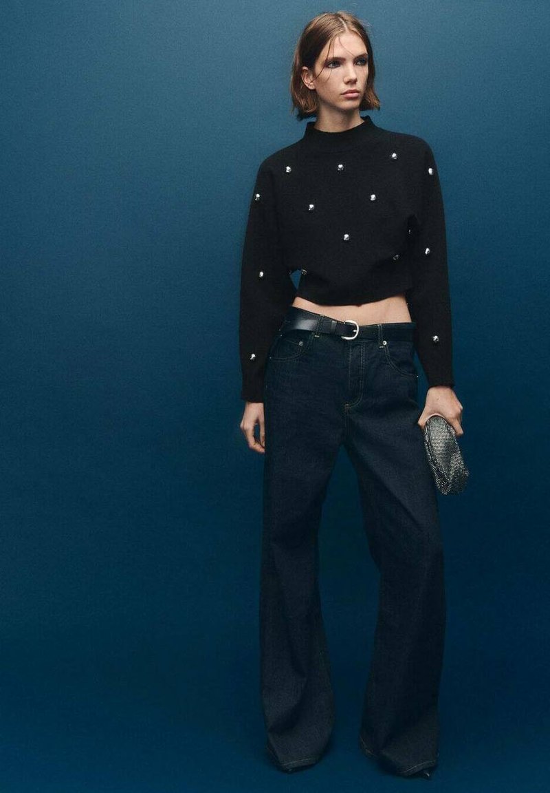 Pullover nero cropped con perline argentate, abbinato a jeans in denim scuri a vita alta. La modella tiene una clutch metallica, in piedi contro uno sfondo blu.