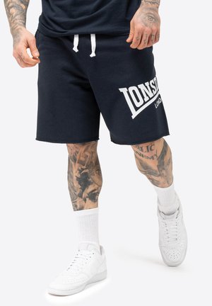 Marineblauwe katoenen shorts met een witte logo-opdruk, elastische tailleband met trekkoord en een relaxed fit, gecombineerd met witte sneakers en sokken.