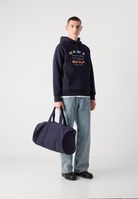 Borsa duffel in canvas blu navy con manici doppi, chiusura zip e logo in rilievo. Il modello indossa un hoodie blu navy abbinato e jeans azzurri.