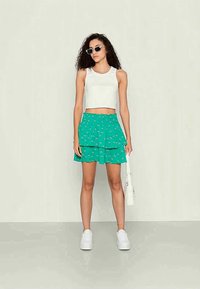 Top cropped branco, saia em camadas com padrão floral verde, sapatilhas brancas e óculos de sol pretos. Design minimalista, estilo casual.