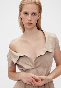 Beige knappklänning med lös krage, korta ärmar och kroppsnära överdel. Mjuk textur, minimal detaljering och avslappnad silhuett.