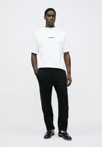 Λευκό κοντομάνικο μπλουζάκι με μαύρο λογότυπο, συνδυασμένο με μαύρο φόρμα. Το σύνολο διαθέτει άνετη εφαρμογή και αίσθηση ομαλής υφής.