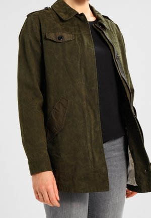 Persona vistiendo una chaqueta de patrón de camuflaje verde oscuro sin cerrar sobre una camisa negra y vaqueros grises, de pie frente a un fondo neutro.